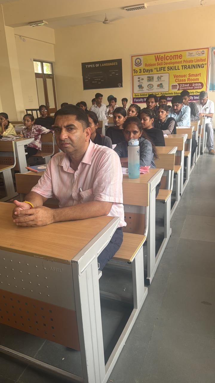 A faculty member seated at a classroom desk with students seated behind him.